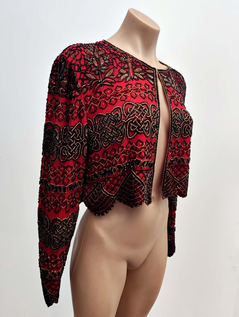 May include: A red, open-front jacket with intricate black and gold beadwork. The jacket features long sleeves and a scalloped hem. The design includes floral and geometric patterns, creating a formal and ornate appearance. The jacket is displayed on a mannequin.