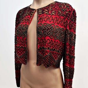 May include: Red bolero jacket with intricate black and gold beadwork. The open-front jacket features long sleeves and a scalloped hem. The design includes floral and geometric patterns, creating a decorative and elegant look.