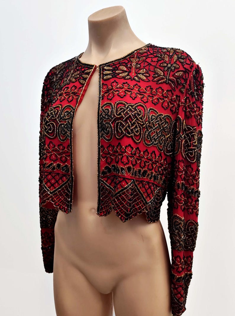 May include: Red bolero jacket with intricate black and gold beadwork. The open-front jacket features long sleeves and a scalloped hem. The design includes floral and geometric patterns, creating a decorative and elegant look.