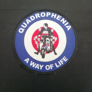 May include: A blue and white circular patch with the text "Quadrophenia A Way of Life" around the edge. The centre of the patch features a red circle with a man riding a scooter.