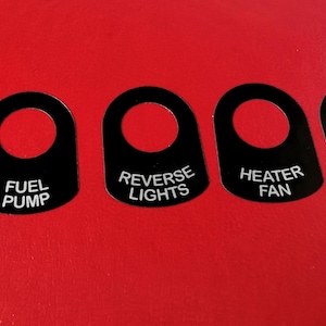 Pode incluir: Cinco tampas de interruptor de plástico preto com letras brancas. As tampas são etiquetadas como "Luzes interiores", "Bomba de combustível", "Luzes de marcha à ré", "Ventilador de aquecimento" e "Sobressalente".