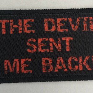 Op de afbeelding: Zwarte stoffen patch met rode tekst die "THE DEVIL SENT ME BACK!" luidt.