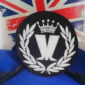 May include: A black circular patch featuring a white crown, the letter "V", and laurel leaves. The patch is set against a backdrop of a distressed Union Jack flag and a blue surface.