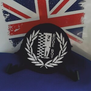 May include: Black patch with the words "RUDE BOY" in white, surrounded by a white laurel wreath. The patch features a checkered design and a silhouette. The background includes a Union Jack flag.