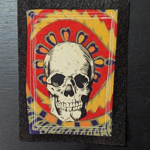 May include: A rectangular fabric patch featuring a black and white human skull illustration centered on a vibrant background of red, yellow, and purple geometric patterns. The patch is bordered by a black felt frame with white stitched detailing.