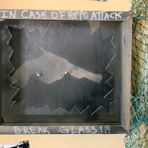 May include: A framed sign with the text "IN CASE OF HIPPO ATTACK BREAK GLASS!" written in white chalk on a black background. The sign has a jagged edge around the glass and two small gold screws.