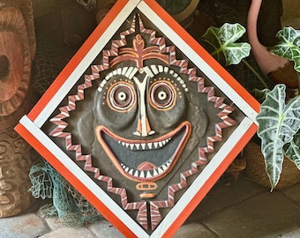May include: A diamond-shaped wall plaque featuring a dark gray sculpted face with wide eyes, a broad smiling mouth, and white teeth. The plaque is framed by a border of alternating orange and white painted wood segments with a zigzag edge.