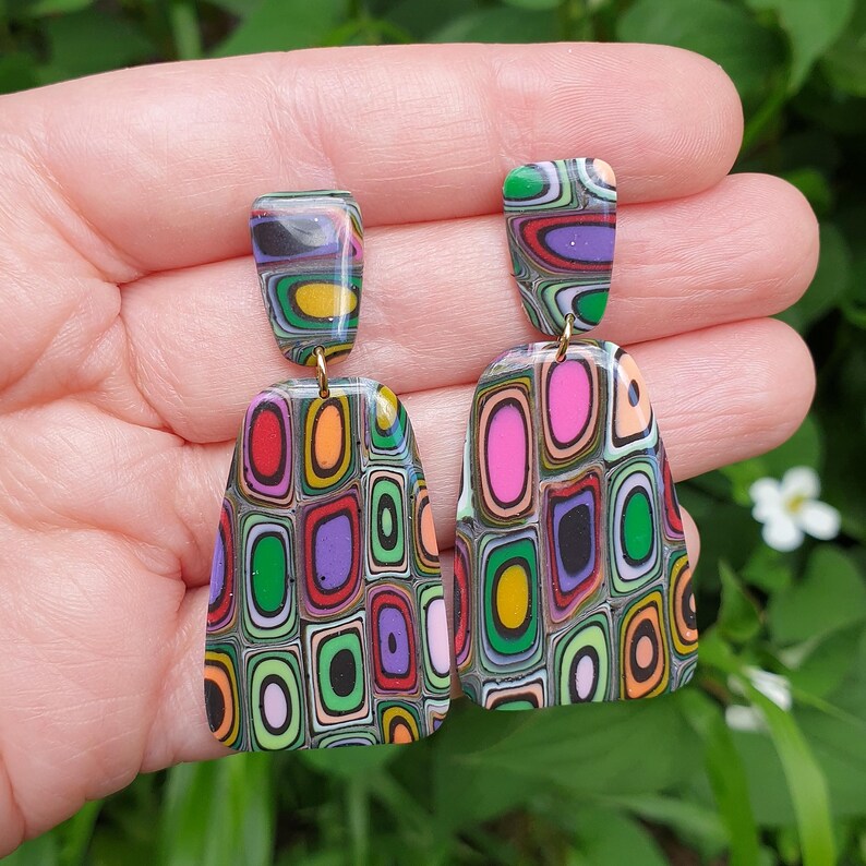 May include: A pair of dangle earrings featuring a multi-colored, layered geometric pattern of nested rectangles in shades of pink, purple, green, orange, yellow, and black. The earrings consist of a small trapezoidal top piece and a larger, wider base.