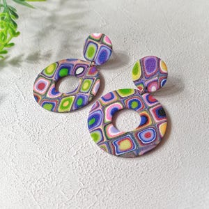 May include: A pair of dangle earrings featuring a small circular stud connected to a larger circular hoop. The earrings display a vibrant, multi-colored geometric pattern of squares with rounded corners in shades of purple, green, blue, pink, and yellow.