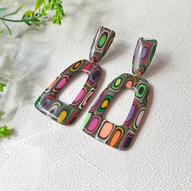 May include: A pair of dangle earrings featuring a glossy, multi-colored layered pattern. Each earring consists of a small rectangular top piece connected to a larger, open-frame trapezoidal bottom piece, showcasing concentric rings of pink, green, orange, and purple.