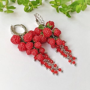 Op de afbeelding: Een paar oorhangers met een cluster van rode polymer clay frambozen die hangen aan een zilveren hoepel met kristal accenten. De oorhangers hebben een hangende ketting van rode kralen.