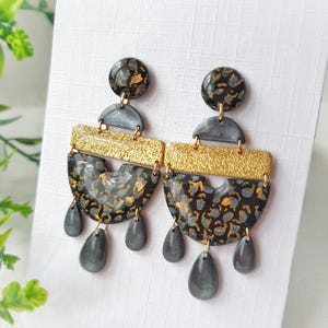 May include: A pair of handmade earrings with a unique design. The earrings feature a black and gold speckled pattern, with gold accents and gray teardrop-shaped dangles. The earrings have a post back.