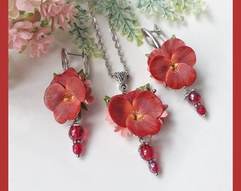 Peut inclure: Un collier avec pendentif en forme de fleur rouge et des boucles d'oreilles assorties. Le pendentif et les boucles d'oreilles présentent une fleur rouge avec un centre rose et sont rehaussés de perles de verre rouges.