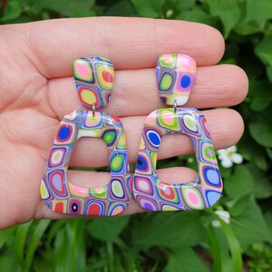 May include: A pair of dangle earrings featuring a multi-colored, abstract geometric pattern in shades of pink, blue, green, yellow, and red. Each earring consists of a small, rounded top piece connected to a larger, open trapezoidal bottom frame.