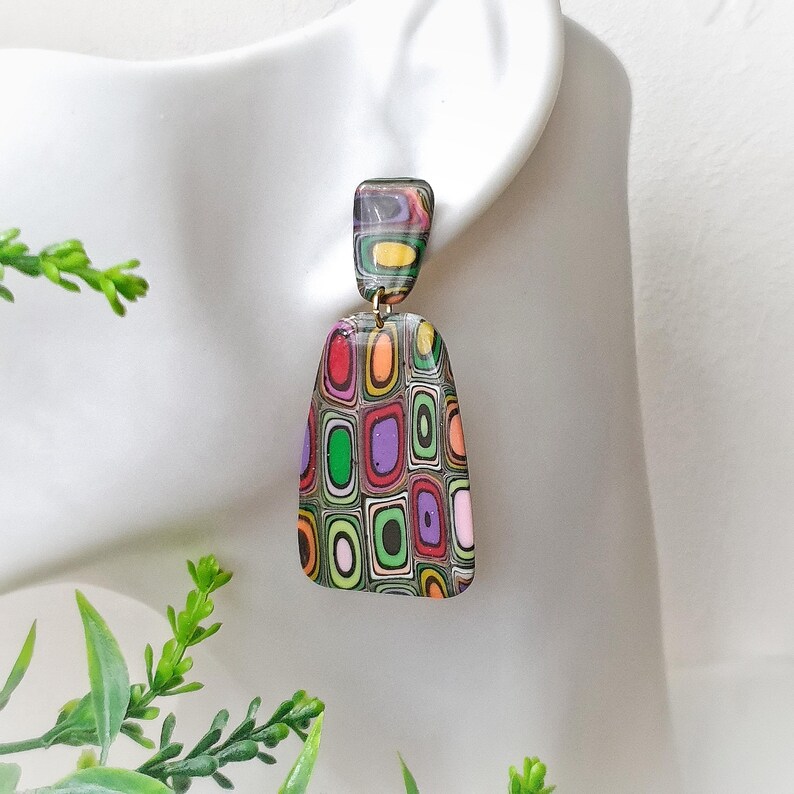 May include: Dangle earrings featuring a two-part design with a small rectangular top piece and a larger trapezoidal bottom piece. Both components display a multi-colored pattern of concentric rounded rectangles in red, orange, green, and purple.