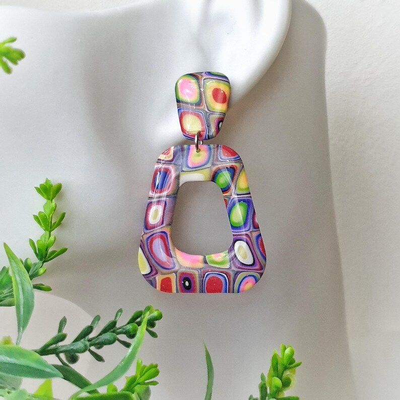 May include: A dangle earring featuring a small rounded rectangular top piece connected to a larger open trapezoidal bottom piece. Both components display a vibrant, multi-colored pattern of small, rounded squares in shades of red, blue, green, yellow, and purple.