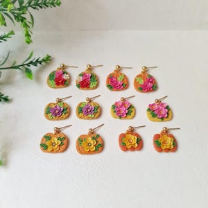 May include: Twelve pumpkin-shaped stud earrings arranged in three rows. Each orange pumpkin features a unique arrangement of colorful sculpted flowers and green leaves. The earrings have gold-toned metal posts and small ball-shaped backings.