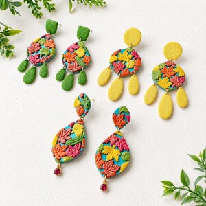 May include: Three pairs of dangle earrings featuring a textured teal base with colorful embossed maple leaves in red, orange, yellow, and green. One pair has green accents, one has yellow accents, and one has small red crystals at the bottom.