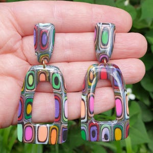 May include: A pair of dangle earrings featuring a multi-colored, layered geometric pattern. Each earring consists of a small rectangular stud connected to a larger, open-frame trapezoidal drop, both displaying vibrant concentric rectangular shapes.
