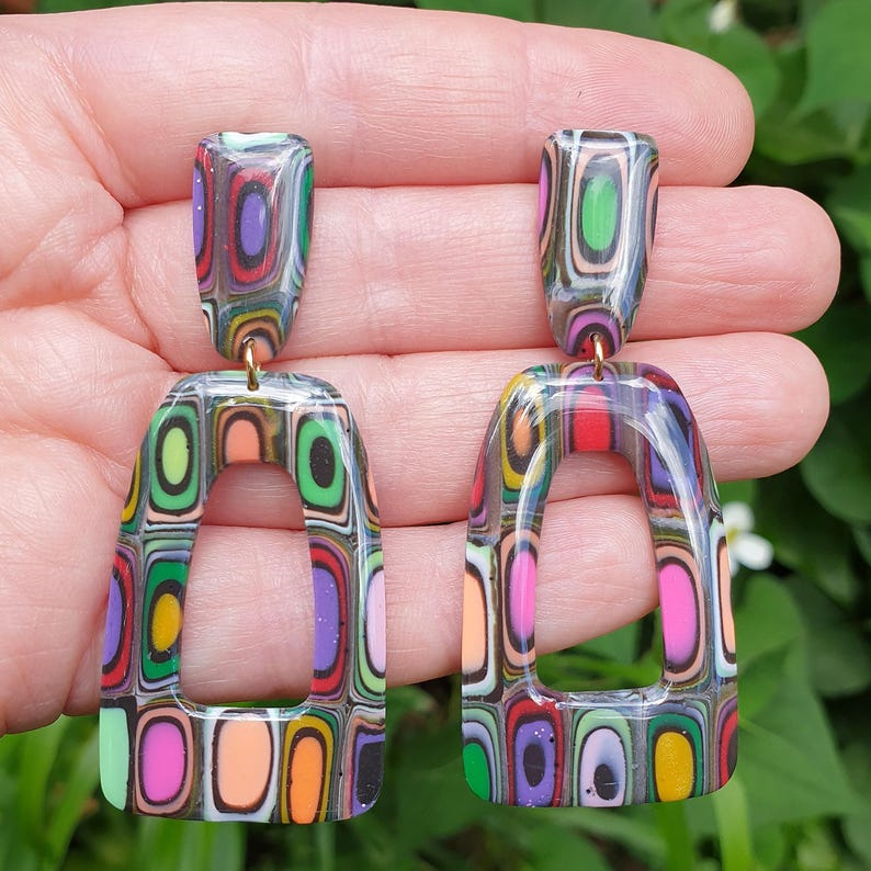 May include: A pair of dangle earrings featuring a multi-colored, layered geometric pattern. Each earring consists of a small rectangular stud connected to a larger, open-frame trapezoidal drop, both displaying vibrant concentric rectangular shapes.