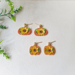 May include: Two pairs of dangle earrings featuring orange pumpkin shapes adorned with yellow sunflowers and green leaves. Each earring has a gold-toned metal post and backing, showcasing a detailed floral design centered on the pumpkin surface.