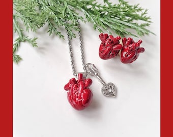 May include: A silver necklace with a red anatomical heart pendant and a silver key charm with a heart-shaped hole. The necklace is paired with a set of red anatomical heart earrings.