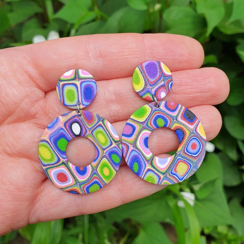 May include: A pair of dangle earrings featuring a small circular stud and a larger circular hoop pendant. Both components display a vibrant, multi-colored geometric pattern of squares and rectangles in shades of blue, green, purple, pink, and yellow.