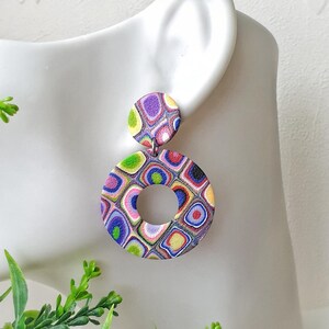 May include: A dangle earring featuring a small circular stud connected to a larger circular pendant with a central cutout. The surface displays a multi-colored pattern of concentric squares in shades of purple, blue, green, pink, and yellow.