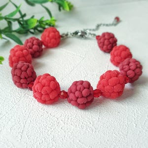 Puede incluir: Una pulsera que presenta múltiples cuentas rojas con forma de frambuesa intercaladas con pequeñas cuentas de cristal rojo translúcido. La pulsera está conectada por una cadena de metal plateado con un cierre de mosquetón y una cuenta roja.