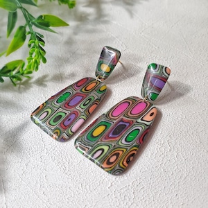 May include: A pair of dangle earrings featuring a multi-colored, layered geometric pattern. Each earring consists of a small rectangular top piece connected to a larger, trapezoidal bottom piece, showcasing concentric rings of pink, green, orange, and purple.