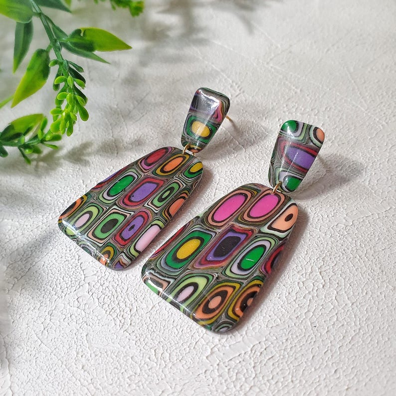 May include: A pair of dangle earrings featuring a multi-colored, layered geometric pattern. Each earring consists of a small rectangular top piece connected to a larger, trapezoidal bottom piece, showcasing concentric rings of pink, green, orange, and purple.