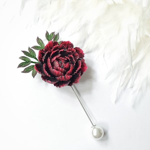 Puede incluir: Un broche de flor de peonía de cuero sintético rojo y negro con hojas verdes y un acento de perla.