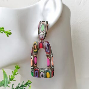 May include: A dangle earring featuring a rectangular top piece and a larger, open-arched bottom piece. Both components display a multi-colored, layered pattern of pink, yellow, green, orange, and purple rectangular shapes set against a dark background.