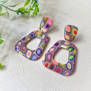 May include: A pair of dangle earrings featuring a multi-colored geometric pattern with shades of pink, blue, green, yellow, and purple. Each earring consists of a small rectangular top piece connected to a larger, open-center trapezoidal shape.