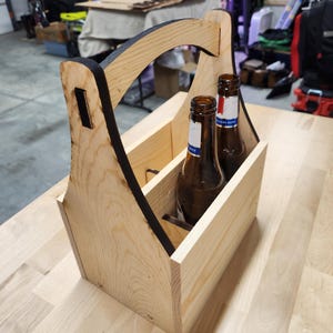 Puede incluir: Un portabotellas de cerveza de madera con asa, que contiene dos botellas de cerveza marrones. El portabotellas es de madera clara con un borde negro en el asa. Las botellas tienen etiquetas blancas y azules.