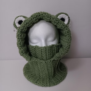 May include: A sage green crochet balaclava featuring two frog-like eyes on top, each with a white and black center. The hood includes a ribbed neck warmer section and silver-toned safety pins attached to the sides of the eyes for decoration.