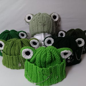 May include: A collection of crochet frog hats in various shades of green. Each hat features large, white-rimmed eyes with black centers and dangling silver chains. The hats are displayed on a white surface.