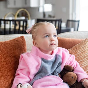 May include: A young child wearing a pink and gray fleece outfit, seated on a couch with orange and patterned pillows. The child is holding a white stuffed animal and a brown stuffed animal.