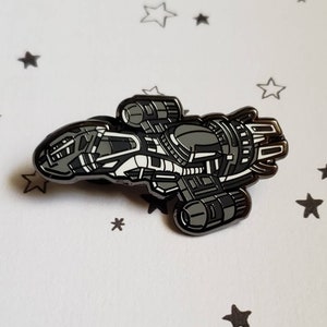 May include: A detailed enamel pin depicting a spaceship in shades of gray and white. The pin features intricate design elements, including windows, engines, and paneling. The pin is set against a white background with scattered star shapes.