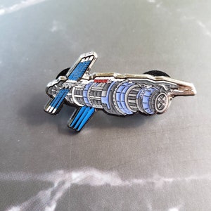 May include: A detailed enamel pin depicting a futuristic spacecraft. The pin features a silver outline with blue and gray internal detailing, and a red accent. The design includes wing-like structures and engine components, suggesting a science fiction theme.