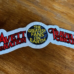 May include: White embroidered patch with the text "Avett Nation" in red and yellow, a black circle with the text "The Greatest Fans On Earth" in yellow, and the text "Traveling Circus" in red and yellow.