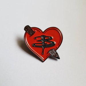 Op de afbeelding: Rode hartvormige emaille pin met zwarte omtrek en zwart 'B' logo. Een bruine pijl doorboort het hart. De pin is een verzamelobject.