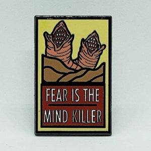 May include: A black and yellow enamel pin with a red banner that reads "FEAR IS THE MIND KILLER". The pin features two cartoon sandworms with open mouths and teeth.