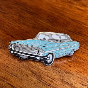 May include: A blue and silver enamel pin of a vintage Ford car with a glittery finish.