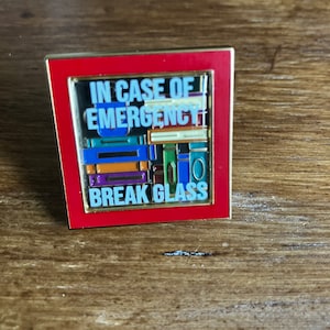 Puede incluir: Un pin de esmalte rojo y dorado con el texto "IN CASE OF EMERGENCY BREAK GLASS" y una pila de libros coloridos detrás del texto.