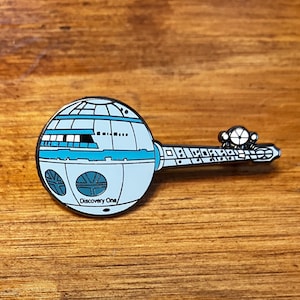 May include: A silver and blue enamel pin of the USS Discovery One spacecraft from the Star Trek television series. The pin features the text "Discovery One" on the side of the spacecraft.