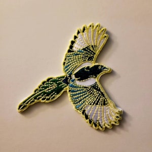 May include: Embroidered patch of a magpie in flight. The bird features a black head and back, white wings with black and teal accents, and a green tail. The patch is outlined in yellow.