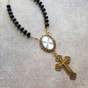 May include: A black beaded necklace with a gold pendant featuring a round medallion with a white, blue, and red flag design and a gold cross.