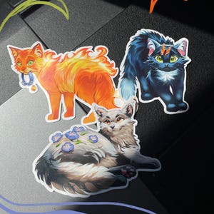 Op de afbeelding: Drie gestanste stickers met ge&iuml;llustreerde katten. E&eacute;n kat is oranje met vlamachtige vacht en houdt een blauwe halsband vast, &eacute;&eacute;n is donkerblauw met een vlinder op zijn kop, en &eacute;&eacute;n is lichtgrijs met blauwe bloemen op zijn rug.