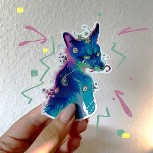 Può includere: Un adesivo di volpe blu e viola con accenti di glitter e un design fantasioso. La volpe ha una caramella a bastoncino in bocca ed è circondata da vortici e stelle colorati.